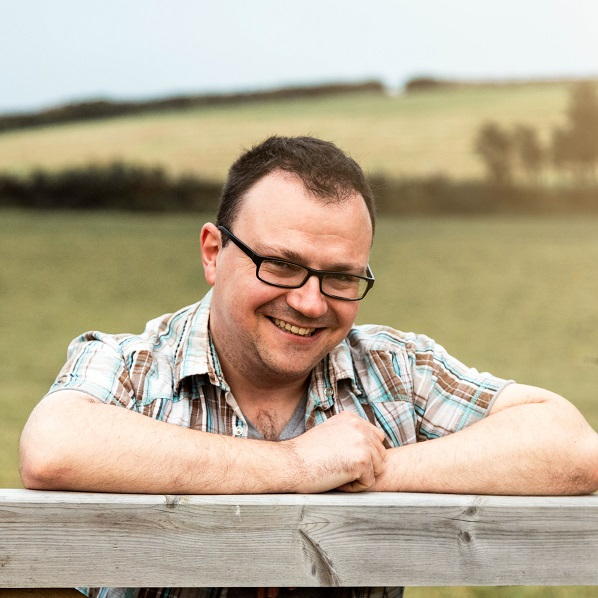 A photo of Dr Daniel Chalk. The photo shows a smiling man with short dark hair and glasses. He is wearing a blue and brown checked shirt and leaning on a fence, with the Cornish countryside in the background.