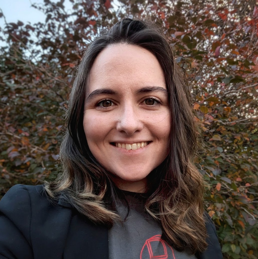 A photo of Sammi Rosser. The photo shows a smiling woman with long wavy brown hair and brown eyes, wearing a grey t-shirt with a red HSMA logo on and a blazer. She is standing in front of a tree with autumnal red, golden and green foliage.